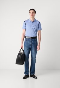 Svetlomodrá polo košeľa s čiernymi akcentmi, modré džínsy, čierny opasok a čierna kožená taška. Model má na sebe čierne topánky a stojí na neutrálnom pozadí.