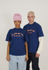Blå bomulds t-shirts med orange grafisk tekst bæres af to personer. Kvinden har en fluffy pink hat, mens manden har en hvid langærmet skjorte under.