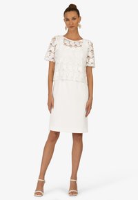 Weißes Kleid mit einem Spitzenoberteil und kurzen Ärmeln, glattem Rock und dekorativem, gewelltem Saum. Trägt dazu bequeme beige Riemchen-High Heels.