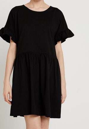 Vrouw die een losse zwarte jurk draagt met korte gerimpelde mouwen en een geplooide taille, staand tegen een effen lichte achtergrond.