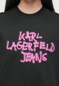 Czarna koszulka z okrągłym dekoltem, z natryskowym różowym napisem "KARL LAGERFELD JEANS" w swobodnej, wyrazistej czcionce.