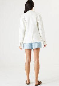 Biała, dopasowana marynarka z długimi rękawami, ozdobiona złotymi guzikami. Składana z jasnoblękitnymi szortami dżinsowymi i brązowymi sandałami.
