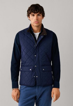 Man draagt een marineblauw doorgestikt vest over een donker overhemd en blauwe broek, staand tegen een effen lichtgrijze achtergrond.