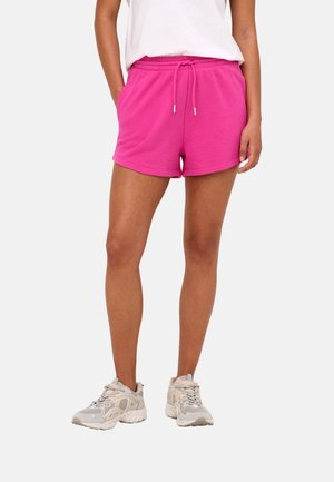 Persoon die felroze short met trekkoord en beige sneakers draagt, staat met handen in de zakken tegen een effen achtergrond.