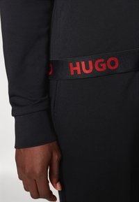 Zwarte sportkleding met een zachte textuur, met een brede tailleband waarop in vette rode letters "HUGO" staat. Close-up van een hand die op de stof rust.