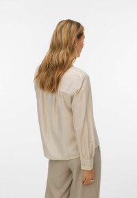 Gaiši bēša blūze ar garām piedurknēm, teksturētu audumu, pogām uz aprocēm un brīvu piegulumu; priekšējā daļā krokojums un smalks mirdzums.