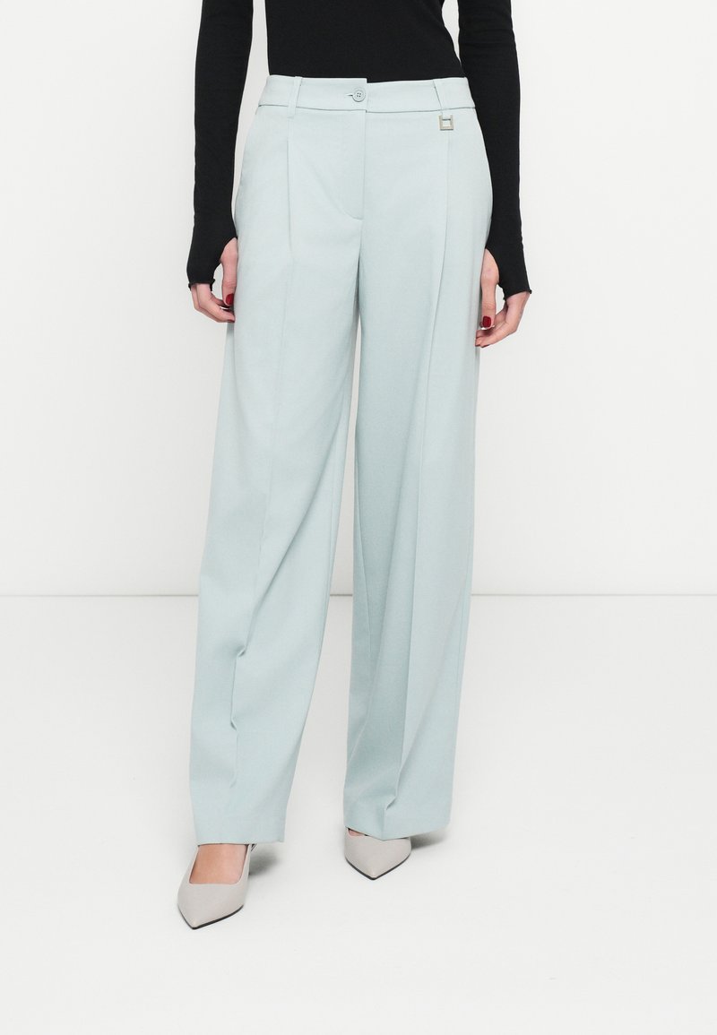 Világoskék, bő szárú nadrág elején redőkkel, gombos záródással és fémrészletekkel. Egy hozzá illő fekete, testhez simuló felsővel és világosszürke magassarkú cipővel viselve.