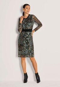 Frau, die in einem transparenten, langärmligen, knielangen Kleid mit Leopardenmuster, schwarzem Taillengürtel und schwarzen Stiefeletten vor einem einfarbigen Hintergrund posiert.