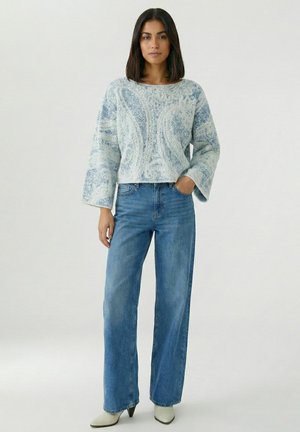 Frau steht und trägt einen hellblauen, gemusterten, flauschigen Pullover, hochtaillierte, weit geschnittene blaue Jeans und hellfarbene, spitz zulaufende Stiefel.