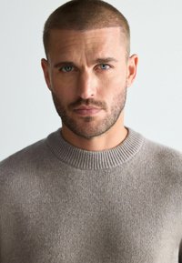 Beżowy sweter dzianinowy z prążkowanym okrągłym dekoldem, z teksturowanym splotem. Model ma krótkie włosy i zarost na twarzy.