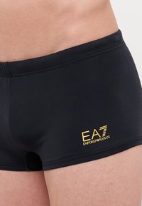 Schwarze Badehosen aus einem glatten Material mit einem klassischen Schnitt. Mit einem goldenen "EA7 Emporio Armani"-Logo auf der linken Vorderseite.