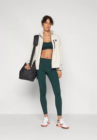 Strój aktywny damski: kremowa polarowa kurtka, ciemnozielone legginsy, czarna torba. Buty sportowe białe z pomarańczowymi akcentami.