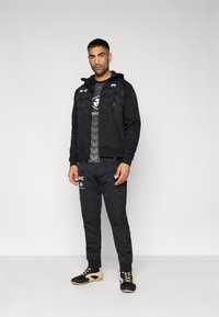 Tenue de sport noire comprenant une veste à fermeture éclair avec un motif texturé et un pantalon assorti. Des chaussures noires et marron complètent le look.