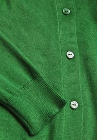 Groene gebreide cardigan met een knoopsluiting aan de voorkant, geribde manchetten en een gladde textuur. Opvallende details zijn de lichtgekleurde knopen.