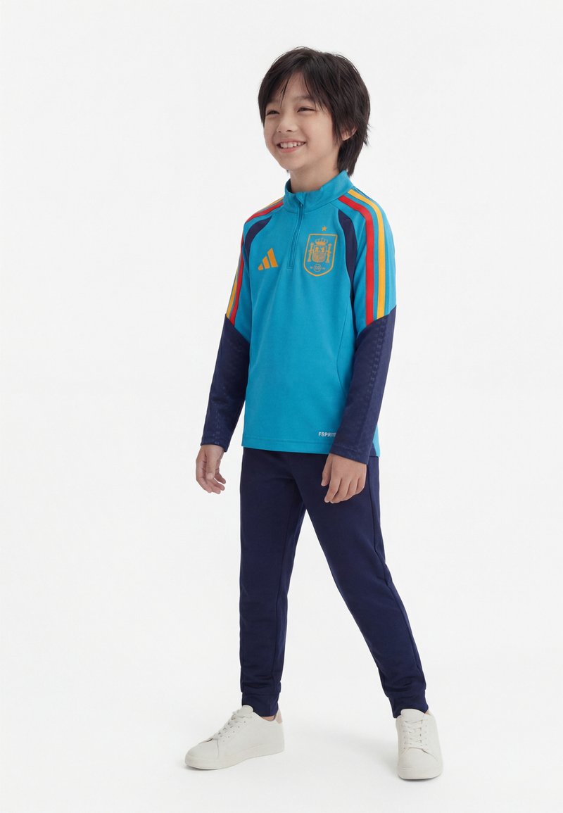 Mosolygó gyermek állva, kék sportdzsekit visel, melyen színes csíkok vannak, és sötét nadrágot egy egyszerű fehér háttér előtt.