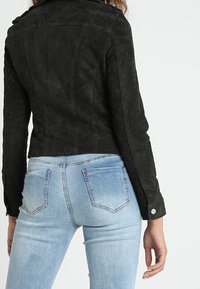 Sort suedejakke med en tætsiddende pasform, synlige sømme og en klippet krave. Båret over lyseblå, udvaskede, lige jeans.