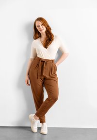 Top cruzado de canalé crema combinado con pantalones marrones de cintura alta que cuentan con un cinturón de lazo, y zapatillas blancas. Fondo neutro.