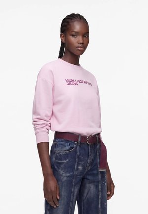 Femme aux cheveux tressés porte un sweat-shirt rose avec le texte "KARL LAGERFELD JEANS" et un jean bleu foncé ample avec une ceinture bordeaux.