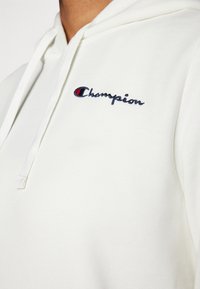 Bílá bavlněná mikina s kapucí a šňůrkou, vyšitým logem "Champion" v námořnické modři a červené na hrudi. Hladký povrch, neformální design.