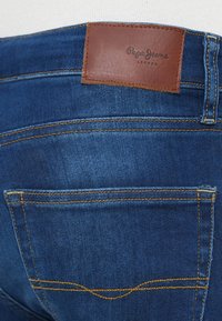 Σκούρο μπλε τζιν υφασμένο με κίτρινη ραφή, με καφέ δερμάτινη ετικέτα που φέρει την επιγραφή "Pepe Jeans London" στη μέση του παντελονιού.