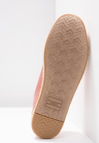 Semelle d'une chaussure TOMS rose avec motif en chevrons et talon en caoutchouc, positionnée à la verticale contre un mur blanc d'angle.