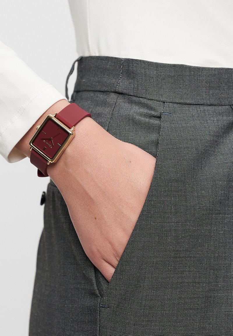 Red square watch with gold accents, plastic strap, worn on a hand. Gray textured pants with a side pocket visible in the image.