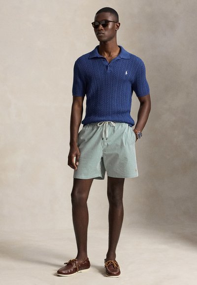 Homme portant des lunettes de soleil foncées, un polo en maille bleu à manches courtes, un short rayé avec cordon et des chaussures bateau marron, debout avec une main dans la poche.