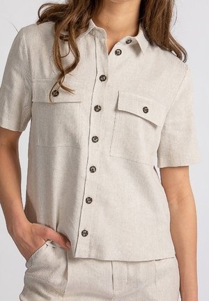 Frau trägt beige kurzärmeliges Hemd mit Knöpfen und zwei aufgesetzten Taschen vorne sowie passende beige Hose, Hand in der Tasche.