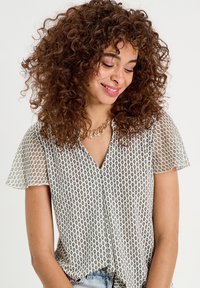 Blouse en tissu léger avec un col en V, des manches courtes et flottantes, et un motif géométrique en noir et blanc. Accentuée avec un collier.