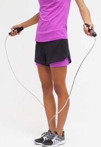 Lila Sportoberteil, schwarze Shorts mit Netzakzenten, grau-schwarze Sneakers und ein Springseil, das in beiden Händen gehalten wird.