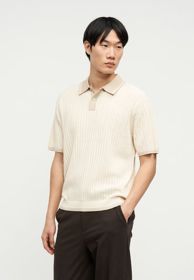 Homme portant un polo en maille à manches courtes crème et un pantalon sombre, debout avec une main dans la poche, regardant légèrement sur le côté.