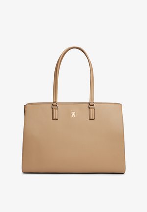 Bēšas ādas soma ar divām siksnām un mazu metāla logotipu, kas novietots centrā zem siksnām.