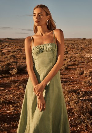 Robe maxi en lin vert avec un corsage froncé, des bretelles fines et une silhouette fluide. Contexte naturel avec des couleurs terreuses en arrière-plan.