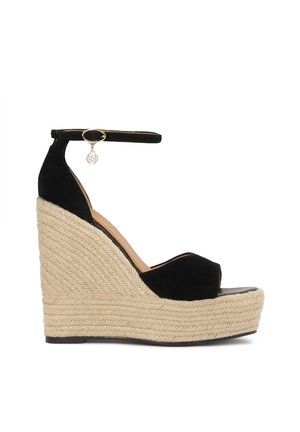 Sandale compensée en daim noir avec une plateforme en jute tissée, une bride de cheville avec un charme en ton doré et un design à bout ouvert.