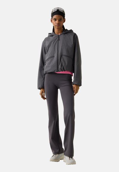 Kobieta stojąca, ubrana w czarną kurtkę z kapturem, ciemne legginsy, białe trampki i czarne okulary przeciwsłoneczne z daszkiem na głowie na białym tle.