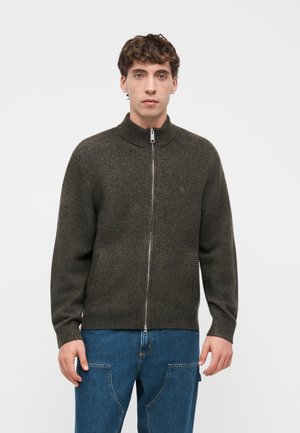 Junger Mann trägt einen dunkelolivfarbenen Wollpullover mit Reißverschluss und blaue Jeans, steht vor einem schlichten hellen Hintergrund.