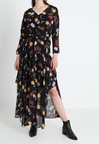 Czarna maxi sukienka w kwiaty z długimi rękawami. Posiada dekolt w serek, warstwowy krój oraz żywe kwiatowe wzory w odcieniach żółtego, czerwonego i fioletowego. Noszona z botkami za kostkę.