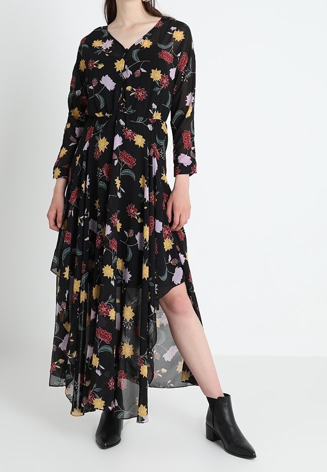 Zwarte maxi-jurk met lange mouwen en bloemenpatroon. Heeft een V-hals, gelaagd ontwerp en levendige gele, rode en paarse bloemenpatronen. G gedragen met enkellaarsjes.