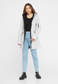 Kobieta w jasnoszarym płaszczu z kapturem, noszącym czarną bluzkę, jasnoniebieskie dżinsy i czarne sznurowane botki, stojąca na tle białym.