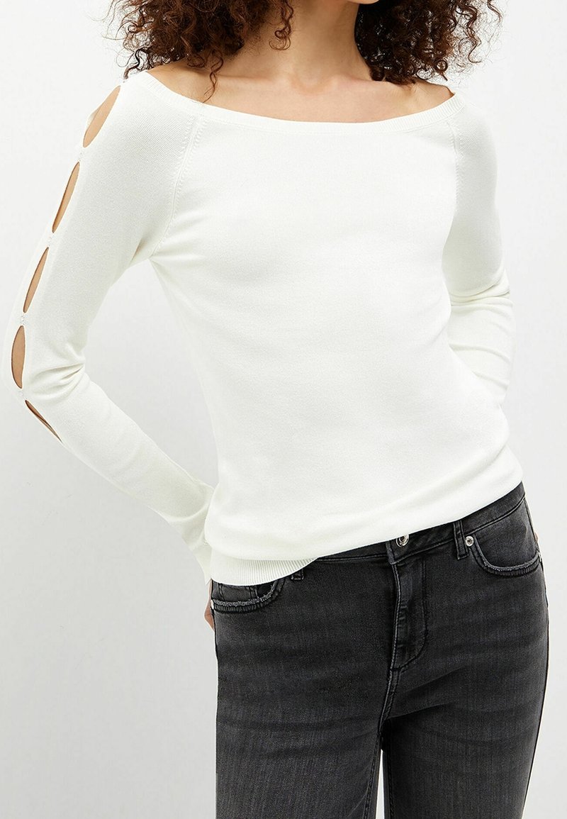 Femme portant un haut blanc à manches longues avec des découpes le long de la manche gauche et un jean noir, debout les mains derrière le dos.