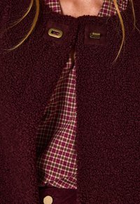 Giacca bordeaux con uno strato esterno testurizzato, dotata di due chiusure in ottone. Fodera in un tessuto a quadri rosso e verde.