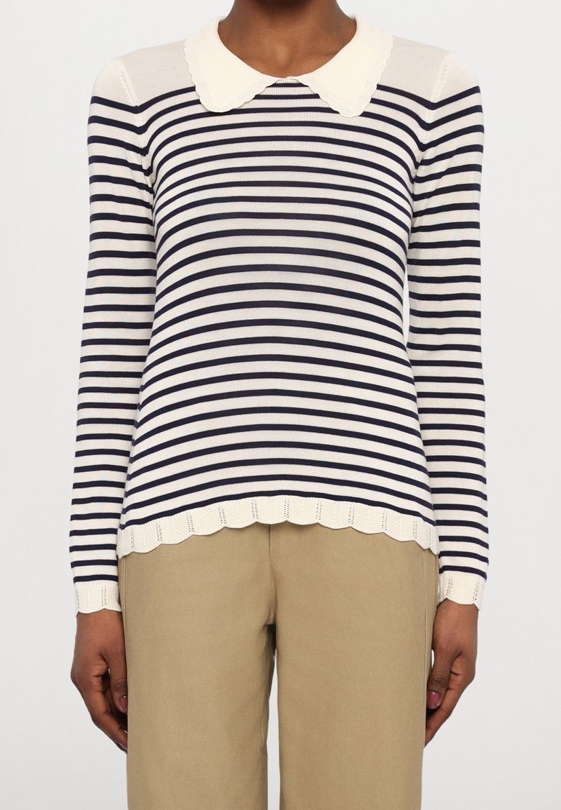Femme portant un pull à manches longues blanc avec des rayures horizontales noires, des poignets et un col à festons, associé à un pantalon beige.