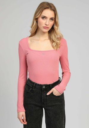 Jeune femme aux longs cheveux blonds portant un haut à manches longues rose et un jean noir taille haute, debout devant un fond clair uni.