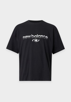 Млад мъж с плитки, носещ черна тениска New Balance и светлосиви панталони на еднороден бял фон.
