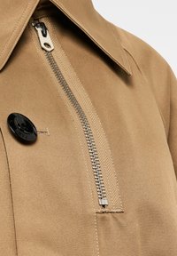 Beżowa kurtka z wyraźnym zamkiem błyskawicznym, dużym czarnym guzikiem oraz gładką fakturą tkaniny. Kołnierz jest szeroki i usztywniony.