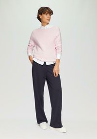 Heller rosa Pullover mit weitem Ausschnitt, kombiniert mit einem weißen Hemd mit Kragen und navyfarbenen weit geschnittenen Hosen. Weiße Sneaker runden das Outfit ab.