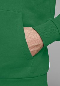 Felpa verde con tasca frontale, polsini a coste e una texture liscia. Dettagli notevoli includono cuciture a vista e un piccolo etichetta di lato.