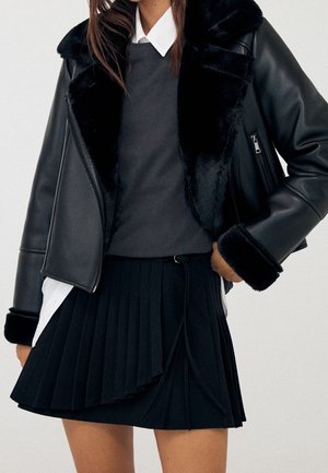 Kobieta w czarnej skórzanej kurtce z futrzanym kołnierzem i mankietami, ciemnoszarym swetrze, białej koszuli oraz czarnej plisowanej spódnicy.