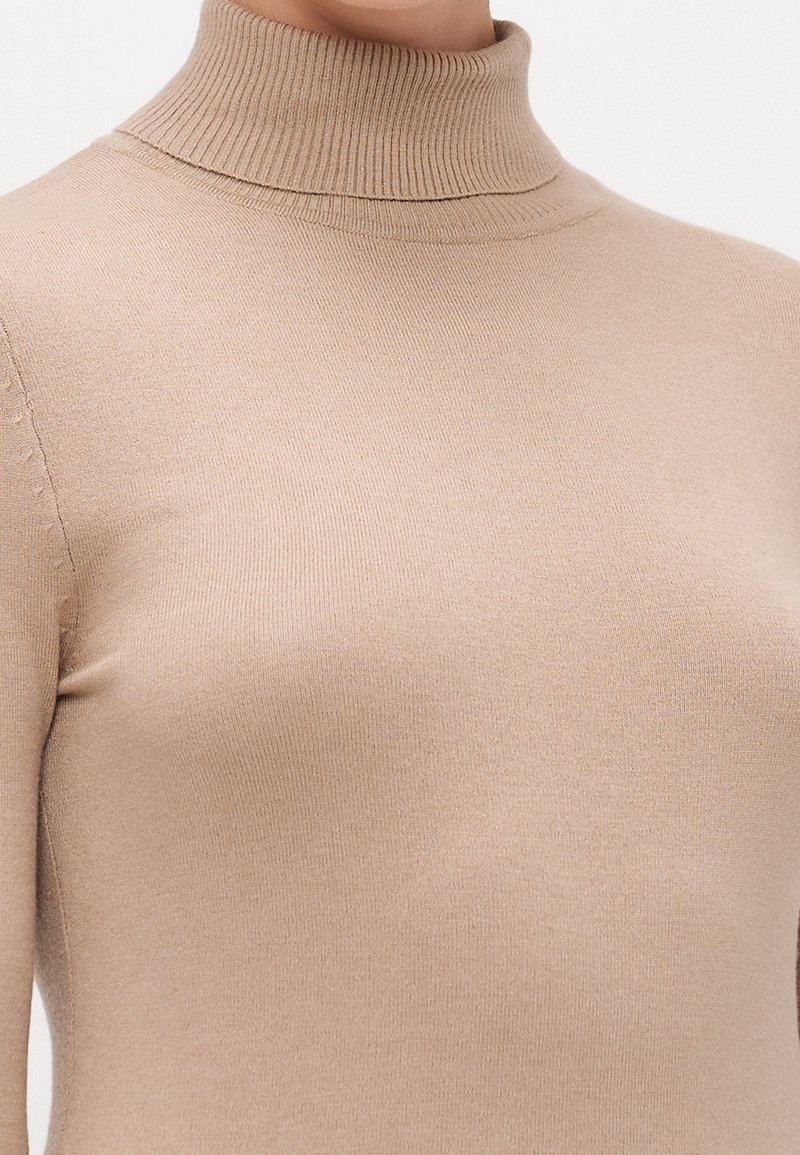 Gros plan d'une personne portant un pull à col roulé côtelé beige à manches longues avec une texture tricotée lisse.