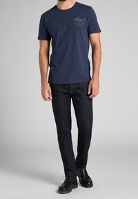 Marineblauwe katoenen t-shirt met een ronde hals en een logo op de linkerborst, gedragen met een donkerdenim jeans en zwarte leren schoenen.